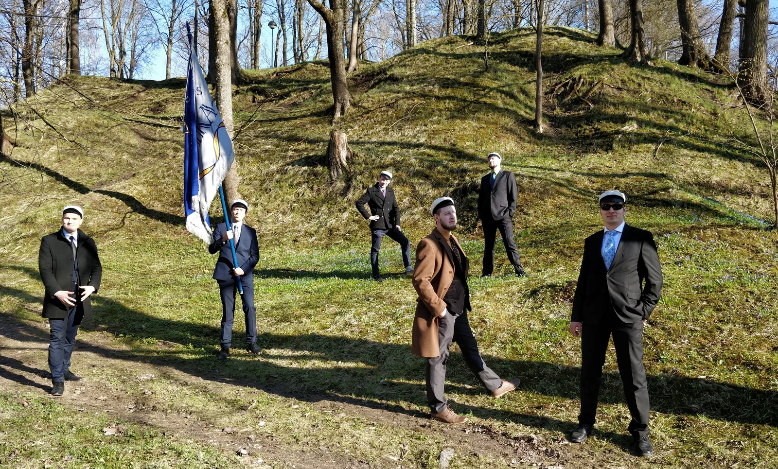 Seitse liiviklast valgetes üliõpilasmütsides ja seltsi lipuga kevadisel mäenõlval puude all, mustvalge foto